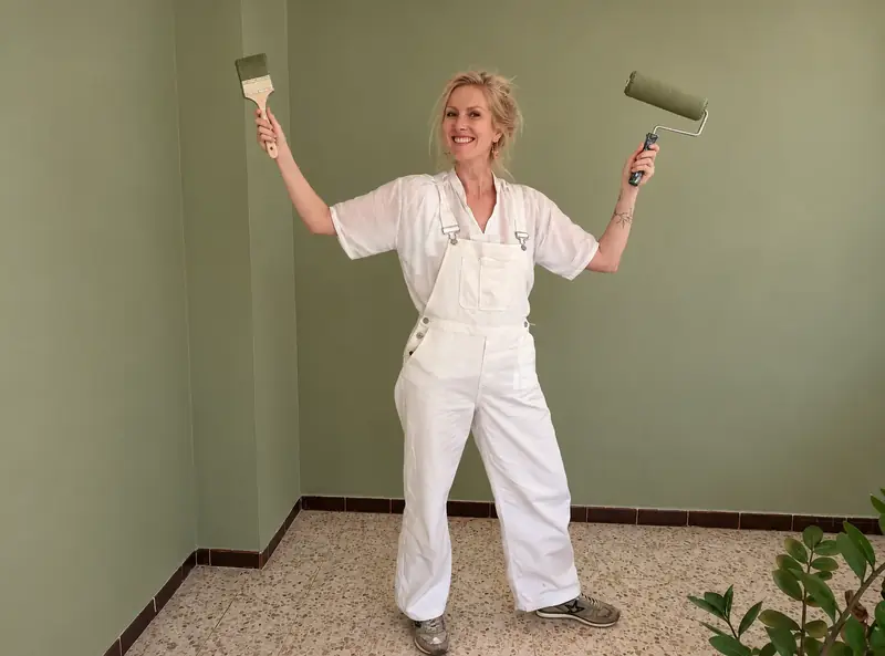 Virginie Georgeault en salopette blanche, rouleau et pinceau en main, dans une pièce peinte en vert sauge